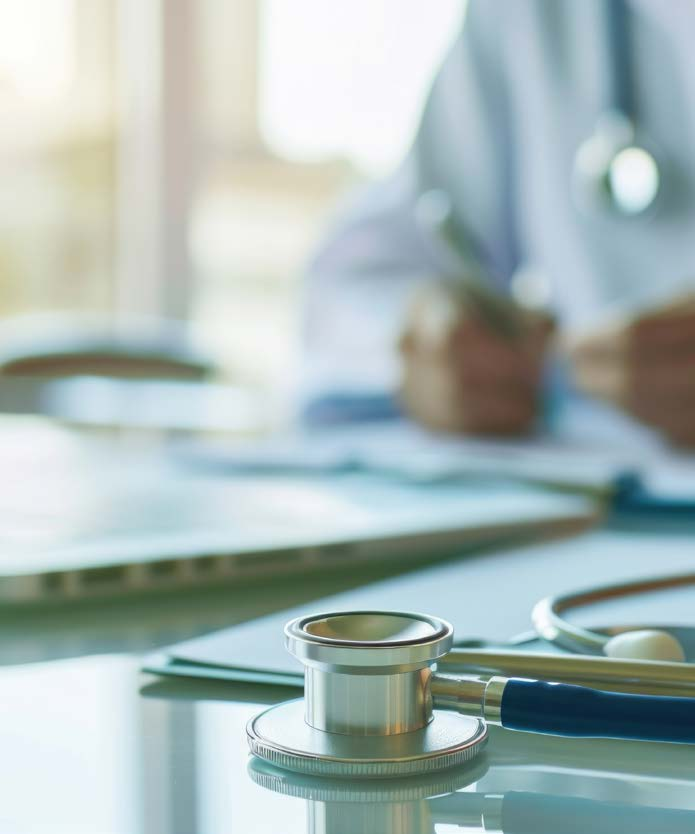 Stetoscopio appoggiato su una scrivania accanto a documenti clinici e a un computer portatile, con un medico in camice sfocato che scrive sullo sfondo